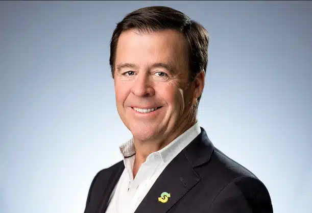 Smiling man in a suit with a pin, against a gray background.