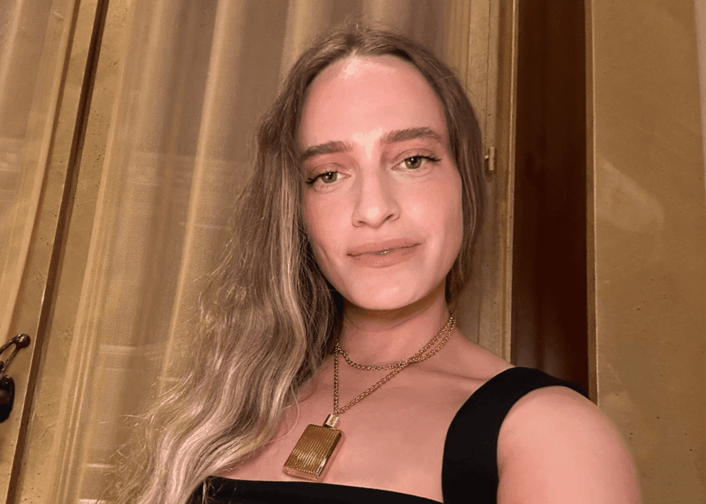 Woman with long hair and makeup, wearing a gold chain necklace, posing indoors.