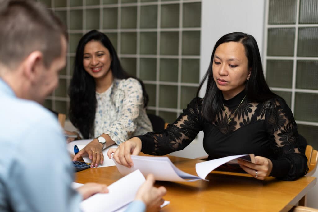 Three people in a meeting, exchanging documents and smiling at a table.