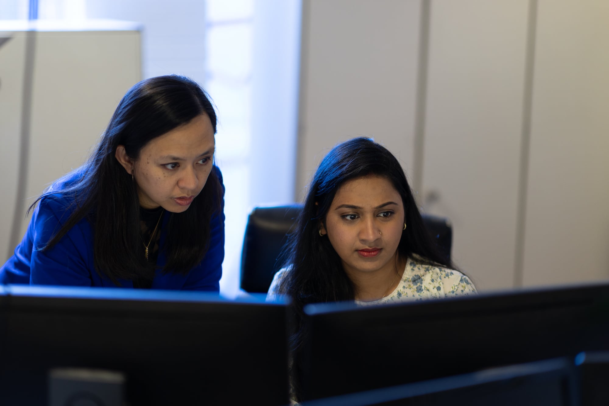 Two women collaborating in an office, focused on computer screens.