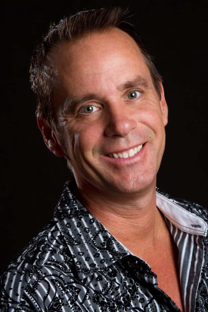 Closeup of a smiling man wearing a patterned shirt against a dark background.